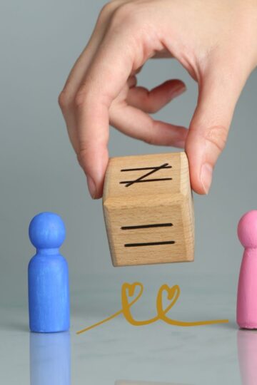 Hand placing a wooden equality block between blue and pink wooden figures, symbolizing gender balance and dismantling patriarchy.